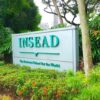 A large sign in a garden setting featuring the word "INSEAD" and the slogan "The Business School for the World." Surrounded by lush greenery and trees.